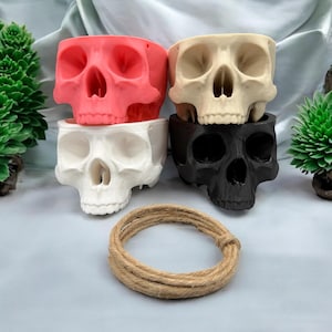 May include: Four skull-shaped planters, stacked in a pyramid. The top planter is beige, the second is pink, the third is white, and the bottom is black.