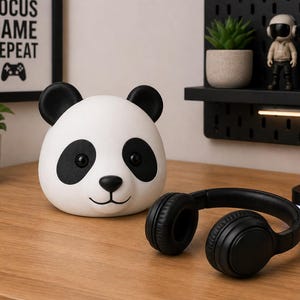May include: A white and black panda-shaped speaker sits on a wooden desk next to a pair of black headphones. A framed print with the words "FOCUS GAME REPEAT" is in the background. A small plant and a figurine are also visible.
