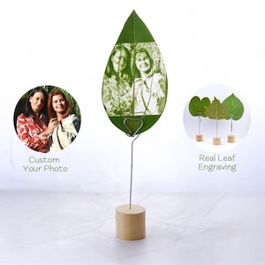 Könnte beinhalten: Ein echtes Blatt mit einem Foto von zwei Personen, das darauf graviert ist. Das Blatt ist auf einem Holzständer mit einem herzförmigen Metallclip montiert. Der Text "Custom Your Photo" und "Real Leaf Engraving" ist unter dem Bild sichtbar.