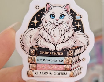 May include: A sticker featuring a white cat with blue eyes, sitting atop a stack of books. The books have the words "CHARMS & CHAPTERS" on the spines. A black arc with stars and a teacup is behind the cat.