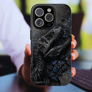 May include: Black phone case featuring a detailed fossilized dinosaur skull design. The case has a dark, textured background with blue accents, and a glossy finish. The phone case is held in a person's hand.