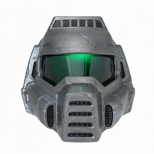 Puede incluir: Un casco gris metálico con un diseño futurista, con una visera verde iluminada. El casco tiene una superficie texturizada y detalles de ventilación visibles, lo que sugiere un tema de ciencia ficción o cosplay.
