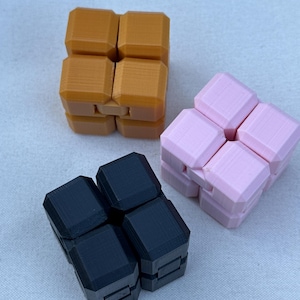 May include: Three fidget cubes are shown. One is orange, one is pink, and the other is black. Each cube is made of eight connected blocks that can be rotated. The cubes are on a light-coloured surface.