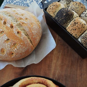 May include: A close-up of two loaves of bread. One loaf is a round, white sourdough bread with a crusty exterior. The other loaf is a dark brown, multi-grain bread with a variety of seeds on top. Both loaves are sitting on a wooden surface.