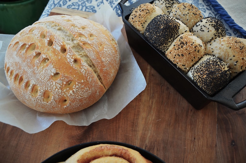 May include: A close-up of two loaves of bread. One loaf is a round, white sourdough bread with a crusty exterior. The other loaf is a dark brown, multi-grain bread with a variety of seeds on top. Both loaves are sitting on a wooden surface.