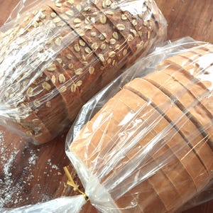 May include: Two loaves of bread, one white and one with oats, are wrapped in clear plastic bags. The loaves are sliced and ready to be eaten.