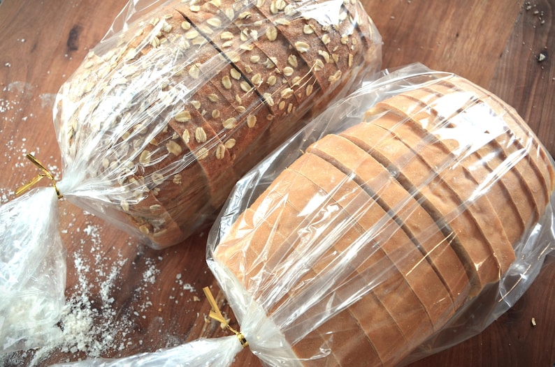May include: Two loaves of bread, one white and one with oats, are wrapped in clear plastic bags. The loaves are sliced and ready to be eaten.