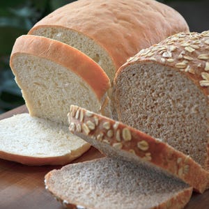 May include: Two loaves of bread, one white and one wheat, sliced on a wooden cutting board. The wheat loaf has a light brown crust and is topped with oats.