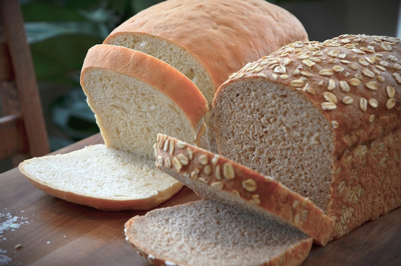 May include: Two loaves of bread, one white and one wheat, sliced on a wooden cutting board. The wheat loaf has a light brown crust and is topped with oats.