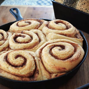 May include: A cast iron skillet filled with six cinnamon rolls, each with a swirl of cinnamon and icing. The rolls are arranged in a circle, with the edges of the rolls overlapping.