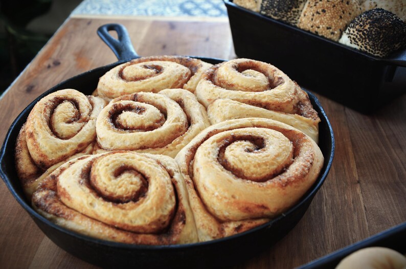 May include: A cast iron skillet filled with six cinnamon rolls, each with a swirl of cinnamon and icing. The rolls are arranged in a circle, with the edges of the rolls overlapping.
