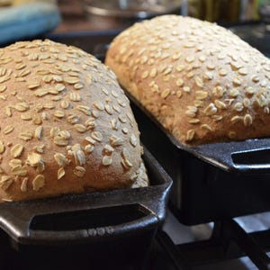 May include: Two loaves of freshly baked bread with a light brown crust and a sprinkle of oats on top. The loaves are in black cast iron bread pans.