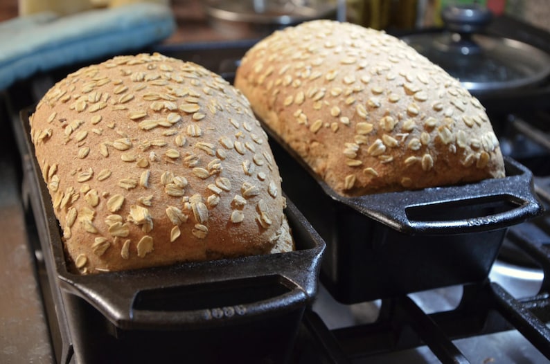 May include: Two loaves of freshly baked bread with a light brown crust and a sprinkle of oats on top. The loaves are in black cast iron bread pans.
