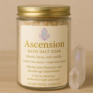 May include: A glass jar with a gold lid filled with bath salts. The label reads "Ascension BATH SALT SOAK" with ingredients listed. Two clear quartz crystals are next to the jar. The jar is on a light brown surface.