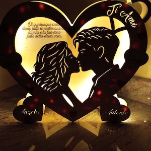 Può includere: Una luce di legno a forma di cuore con la silhouette di una coppia che si bacia. Le parole "Ti Amo" e una citazione in italiano sono visibili. La luce ha luci rosse e i nomi "Angie" e "Antonis" sono visibili.