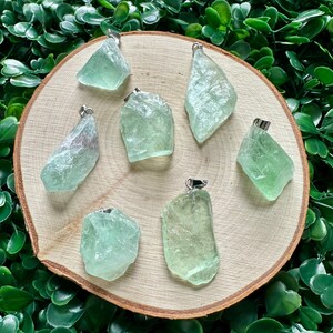 May include: Seven green fluorite crystal pendants with silver bail settings. The crystals are various shapes and sizes and are displayed on a wooden surface.