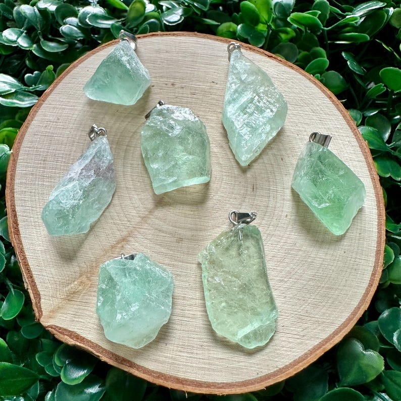 May include: Eight green fluorite crystal pendants with silver bail. The crystals are various shapes and sizes. The pendants are displayed on a wooden surface.