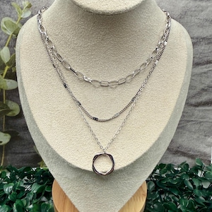 May include: A silver-toned layered necklace displayed on a mannequin. The necklace features three strands: a chain link, a delicate beaded strand, and a pendant with a circular, open design. The mannequin is set against a neutral background with greenery.