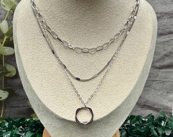 May include: A silver-toned layered necklace displayed on a mannequin. The necklace features three strands: a chain link, a delicate beaded strand, and a pendant with a circular, open design. The mannequin is set against a neutral background with greenery.