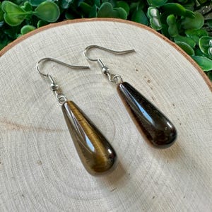 May include: A pair of teardrop-shaped tiger's eye earrings with silver hooks. The earrings are a dark brown colour with gold flecks.