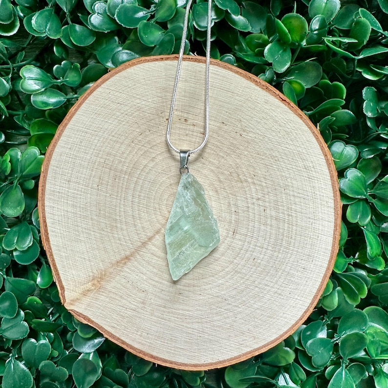 May include: A silver chain necklace with a light green, raw, crystal pendant. The pendant is a natural, irregular shape and has a smooth, polished surface.