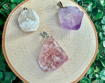 May include: Three raw crystal pendants on a wooden surface. The pendants are attached to silver chains. The crystals are clear, purple, and pink.