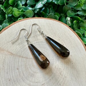 May include: A pair of teardrop-shaped earrings with a dark brown and black wood-like pattern. The earrings are made with silver hooks.