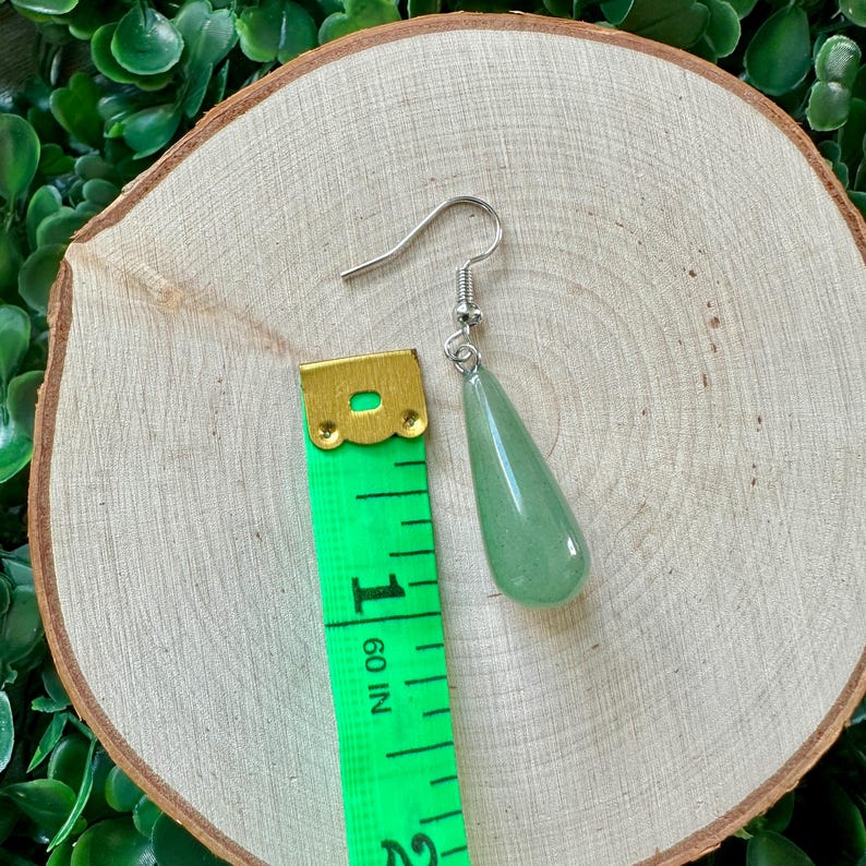 May include: A green teardrop-shaped gemstone earring with a silver hook. The earring is measured against a green measuring tape.