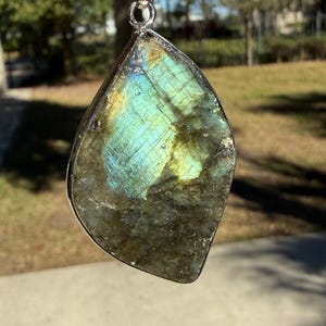 May include: A teardrop-shaped labradorite pendant with a silver-colored metal border. The stone displays iridescent flashes of blue, green, and yellow. The pendant hangs from a silver-colored chain, showcasing natural gemstone jewelry.