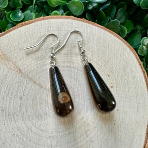 May include: A pair of teardrop-shaped earrings with a black and brown marbled pattern. The earrings are made of resin and have silver hooks.