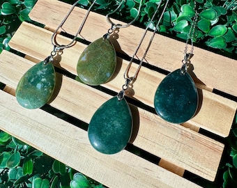 May include: Four green agate stone pendants on silver chains. The stones are teardrop shaped and have a mottled green and white pattern. The pendants are displayed on a wooden surface.