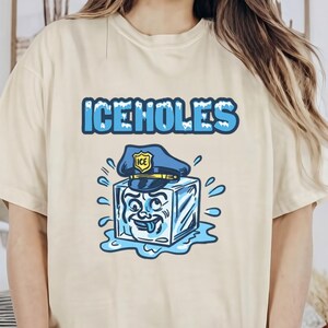 Peut inclure: T-shirt crème avec le texte bleu "ICEHOLES" au-dessus d'un glaçon de dessin animé portant un chapeau de policier. Le glaçon a un visage souriant et est entouré de gouttelettes d'eau. Le t-shirt est de coupe ample.