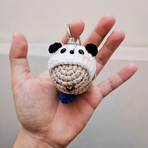 May include: A handmade crochet panda bear keychain. The panda has a white hat with black ears, a beige body, and a small blue pom-pom. The keychain is attached to a silver metal ring.