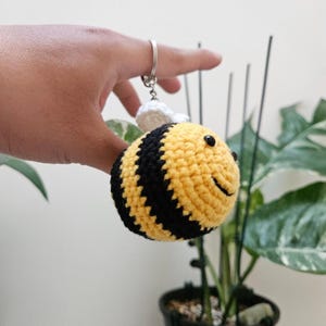May include: A crocheted bee keyring. The bee is yellow with black stripes, small black eyes, and a smiling face. It has white wings and a silver-coloured ring for attaching keys.