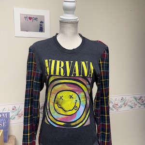 May include: A dark gray long-sleeve shirt with the word "NIRVANA" in yellow block letters above a smiley face design. The sleeves are a plaid pattern in red, blue, and yellow.