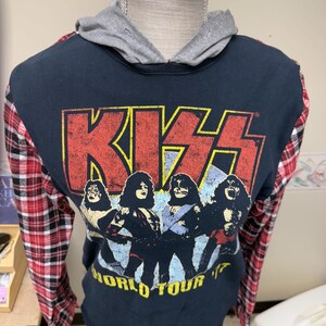 May include: A black hooded sweatshirt featuring a graphic of the band KISS with the text "WORLD TOUR 17". The sweatshirt has red and black plaid sleeves and a gray hood.