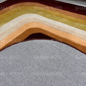 May include: Close-up of a multi-colored rug with curved lines. The rug features shades of brown, beige, and green, with a gray base. The text "SHAMA RUGS" is visible across the top of the rug.