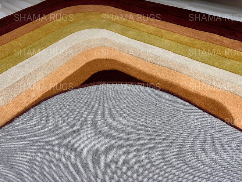 May include: Close-up of a multi-colored rug with curved lines. The rug features shades of brown, beige, and green, with a gray base. The text "SHAMA RUGS" is visible across the top of the rug.
