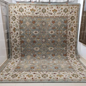 Puede incluir: Una alfombra rectangular grande con un intrincado diseño floral. La alfombra presenta un campo central de color azul grisáceo apagado, bordeado por un borde color crema con motivos florales y de hojas en tonos marrón, dorado y verde. La alfombra se exhibe sobre un suelo claro.