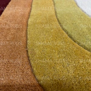 May include: Close-up of a multi-colored rug with a wave-like design. The rug features shades of burgundy, brown, gold, and green. The texture appears soft and plush, with the words "SHAMA RUGS" repeated across the image.