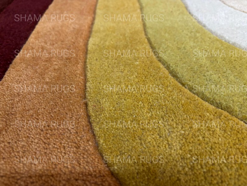 May include: Close-up of a multi-colored rug with a wave-like design. The rug features shades of burgundy, brown, gold, and green. The texture appears soft and plush, with the words "SHAMA RUGS" repeated across the image.
