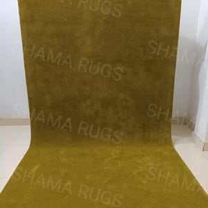 Puede incluir: Una alfombra grande y rectangular de color verde oliva con una textura suave y afelpada. La alfombra est&aacute; sostenida por manos invisibles, mostrando su tama&ntilde;o y el logotipo "SHAMA RUGS". La alfombra parece estar hecha de un material grueso y tejido.