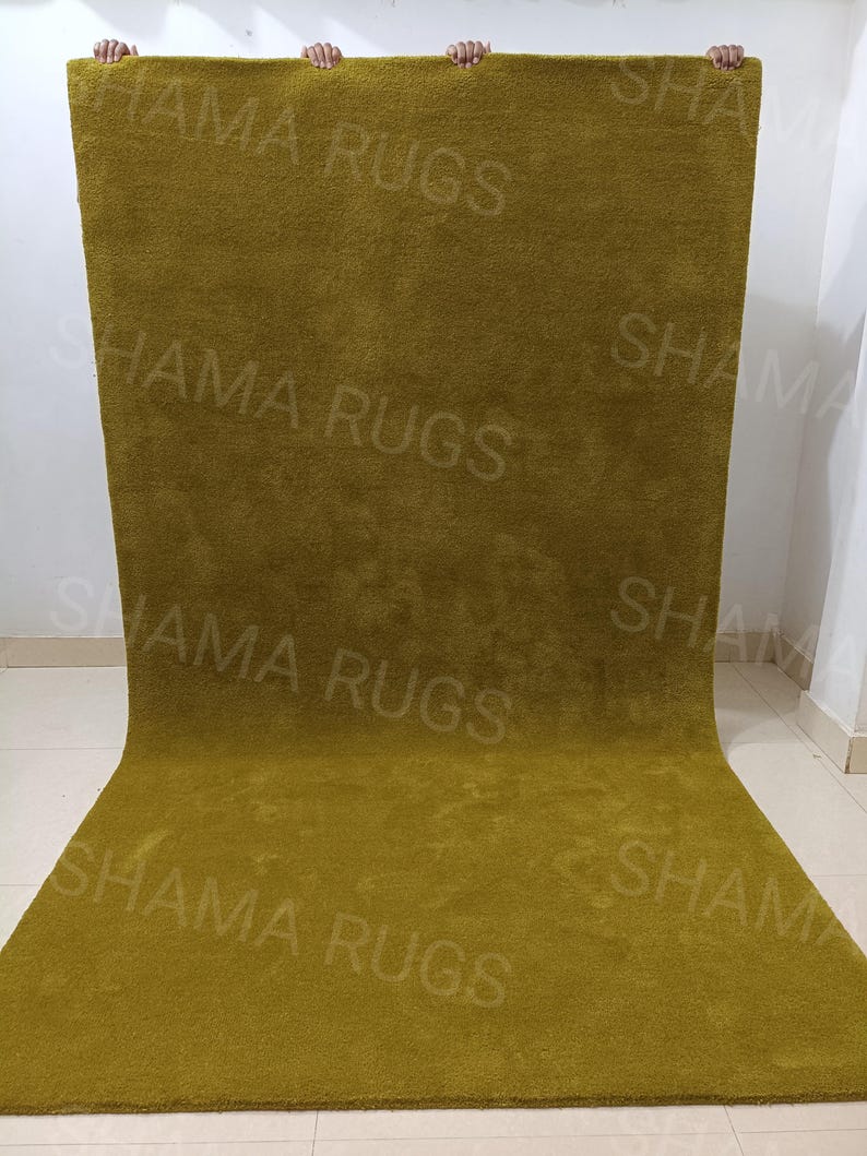Puede incluir: Una alfombra grande y rectangular de color verde oliva con una textura suave y afelpada. La alfombra est&aacute; sostenida por manos invisibles, mostrando su tama&ntilde;o y el logotipo "SHAMA RUGS". La alfombra parece estar hecha de un material grueso y tejido.