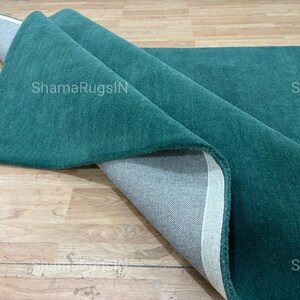 Puede incluir: Una alfombra verde azulado parcialmente enrollada, que revela un reverso gris y un borde blanco. La textura afelpada de la alfombra es visible. El texto "ShamaRugsIN" est&aacute; impreso en la alfombra.
