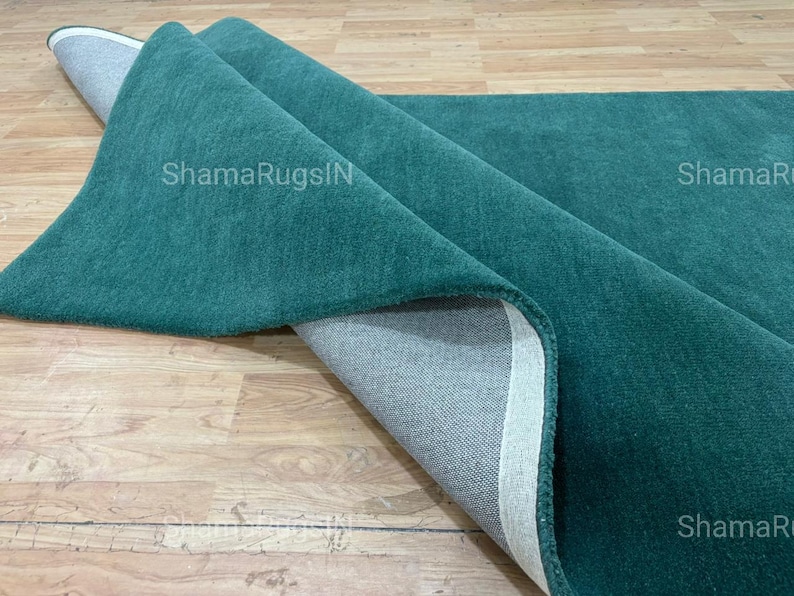 Puede incluir: Una alfombra verde azulado parcialmente enrollada, que revela un reverso gris y un borde blanco. La textura afelpada de la alfombra es visible. El texto "ShamaRugsIN" est&aacute; impreso en la alfombra.