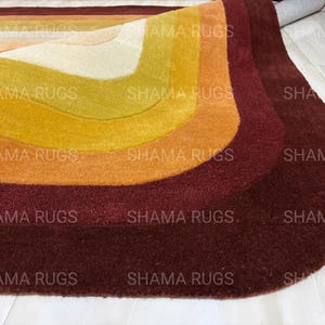 May include: A colorful, retro-style rug with a layered design. The rug features curved bands of color, including cream, yellow, orange, and burgundy. The rug is made of a soft, plush material and is suitable for a living room or bedroom.