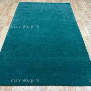 Puede incluir: Una alfombra rectangular de color verde azulado con un dise&ntilde;o de color liso. La alfombra est&aacute; colocada sobre un suelo de madera de color marr&oacute;n claro. El texto "ShamaRugsIN" es visible en la alfombra.