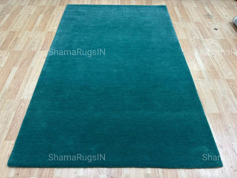 Puede incluir: Una alfombra rectangular de color verde azulado con un dise&ntilde;o de color liso. La alfombra est&aacute; colocada sobre un suelo de madera de color marr&oacute;n claro. El texto "ShamaRugsIN" es visible en la alfombra.