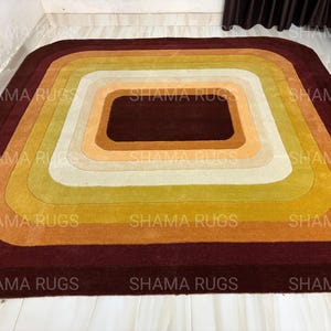 May include: A rectangular rug with a retro design, featuring concentric squares in shades of brown, orange, yellow, and cream. The rug has rounded corners and a dark brown border, creating a warm and inviting aesthetic. The text "SHAMA RUGS" is visible.