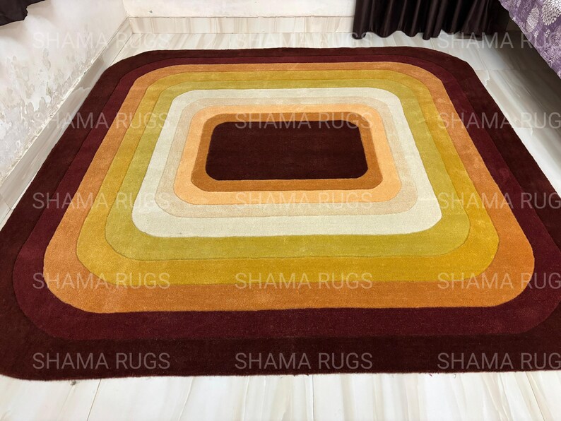May include: A rectangular rug with a retro design, featuring concentric squares in shades of brown, orange, yellow, and cream. The rug has rounded corners and a dark brown border, creating a warm and inviting aesthetic. The text "SHAMA RUGS" is visible.